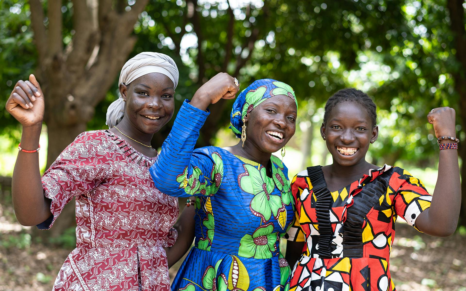 Three African women in colorful traditional dresses raise their fists as a symbol for women's strength and gender equality.