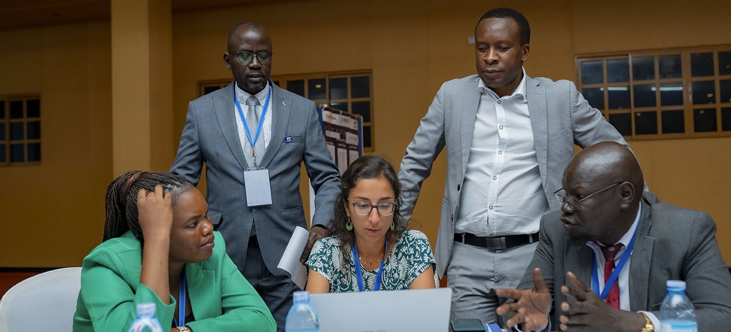 A group of two women and three men having a discussion while looking at a laptop computer in a conference room in Uganda. They are dressed in business attire.