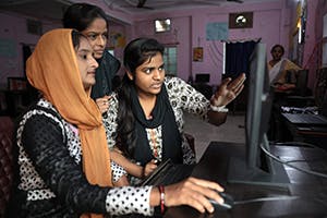 Three young Indian women are discussing information they are looking at on a computer screen.